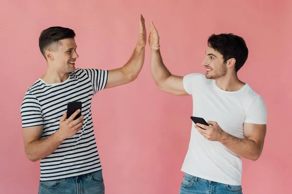 Dos hombres sonrientes usando teléfonos inteligentes y mirándose aislados en rosa - foto de stock
