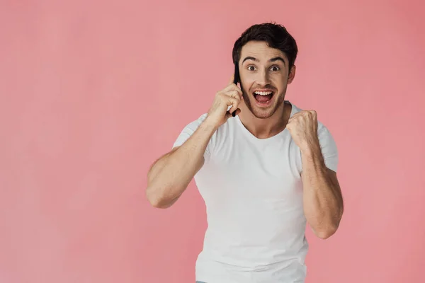 Visão frontal do homem muscular animado em t-shirt branca falando no smartphone e mostrando sim gesto isolado em rosa — Fotografia de Stock