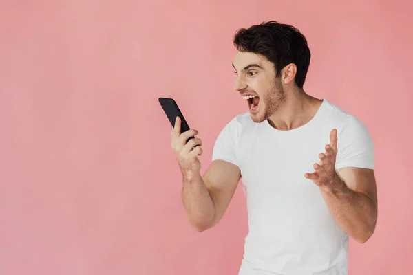 Agresivo musculoso hombre en blanco camiseta sosteniendo teléfono inteligente y gritando aislado en rosa - foto de stock