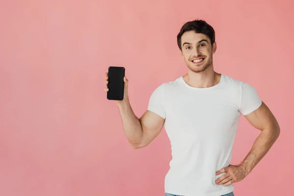Вид спереду сміється м'язистий чоловік у білій футболці, що стоїть з рукою на стегні і тримає смартфон з порожнім екраном ізольовано на рожевому — стокове фото