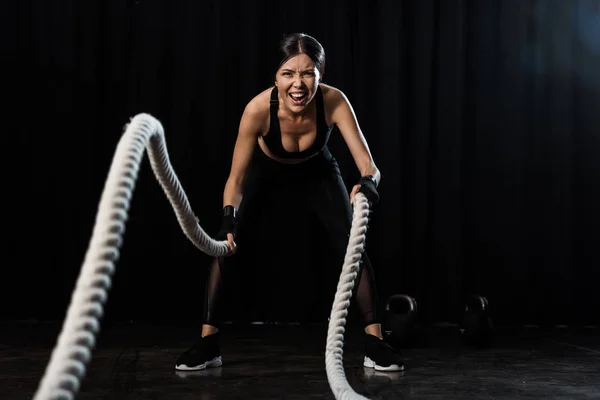 Selective focus of sportswoman exercising with battle ropes on black — Stock Photo