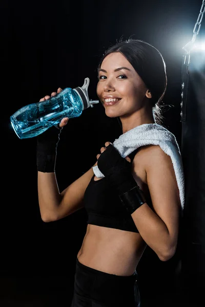 Счастливая девушка, держа в руках спортивную бутылку и улыбаясь изолированно на черном — стоковое фото