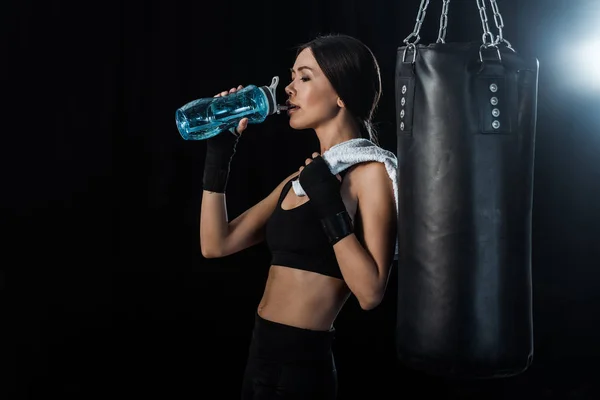 Chica beber agua de deporte botella y de pie cerca de saco de boxeo aislado en negro - foto de stock