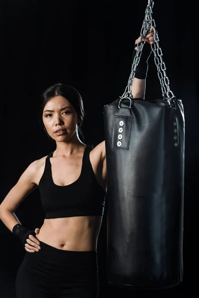 Attractive girl standing with hand in hip near punching bag isolated on black — Stock Photo