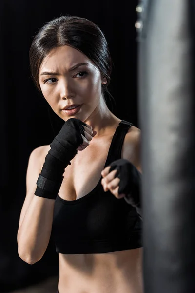 Enfoque selectivo de hermosa chica haciendo ejercicio cerca de saco de boxeo aislado en negro - foto de stock
