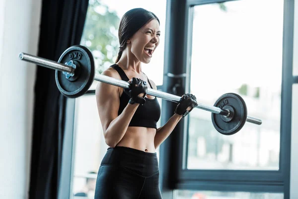 Emotionale junge Frau in Sportbekleidung trainiert mit Langhantel im Fitnessstudio — Stockfoto