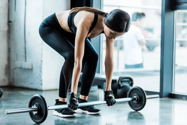 Jeune femme en vêtements de sport prenant lourd haltère dans la salle de gym — Photo de stock