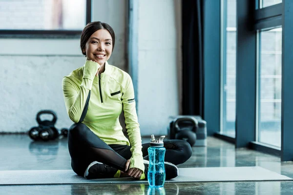 Felice giovane donna seduta sul tappeto fitness vicino bottiglia di sport con acqua — Foto stock