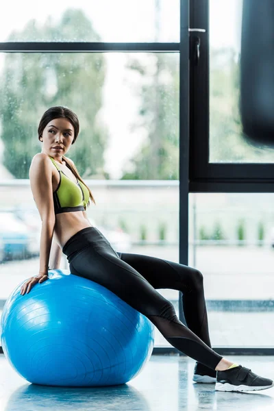 Selektiver Fokus des schönen Mädchens, das auf blauem Fitnessball in der Nähe von Hanteln sitzt — Stockfoto