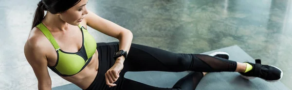 Panoramaaufnahme einer attraktiven Frau, die auf der Fitnessmatte sitzt und auf die Fitnessuhr schaut — Stockfoto