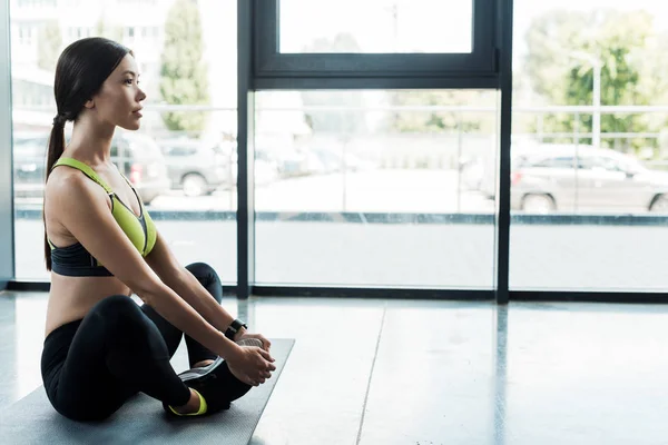 Schöne junge Frau dehnt sich auf Fitnessmatte in der Nähe von Fenstern im Fitnessstudio — Stockfoto