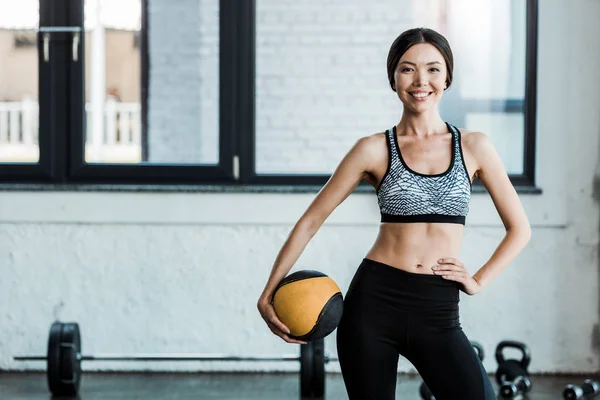 Cheerful sportswoman standing with hand on hip and holding ball — Stock Photo
