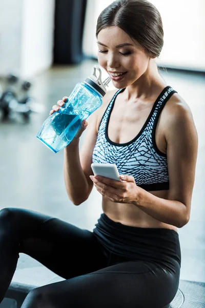 Sportive heureuse utilisant un smartphone et une bouteille de sport avec de l'eau — Photo de stock