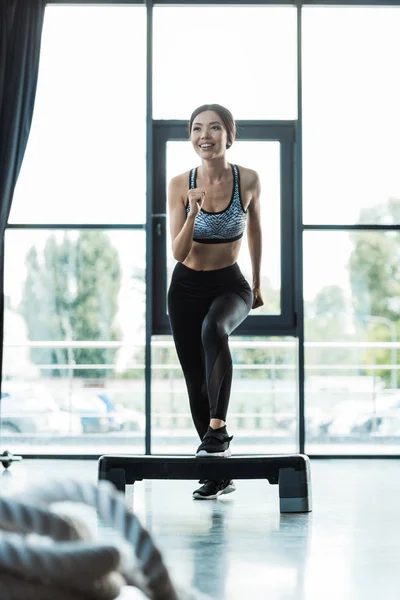 Selective focus of cheerful girl working out on step platform in sports center — Stock Photo