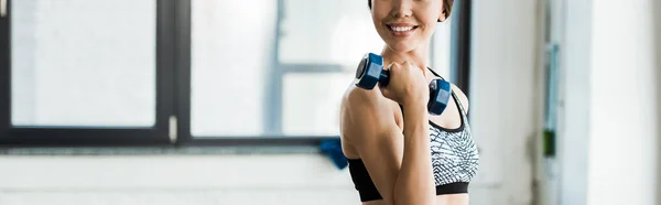 Panoramic shot of cheerful young woman working out with dumbbell and smiling in gym — Stock Photo