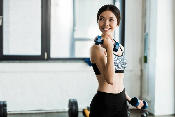 Alegre joven mujer haciendo ejercicio con mancuernas y sonriendo en el gimnasio - foto de stock