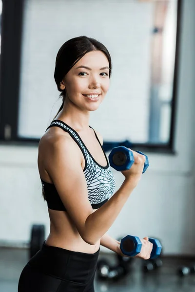 Mujer positiva y deportiva sosteniendo pesas mientras hace ejercicio en el gimnasio - foto de stock