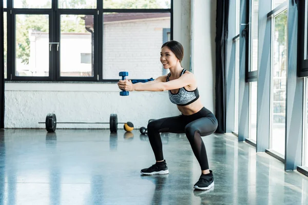 Jovem desportista feliz segurando halteres e fazendo exercício agachamento — Fotografia de Stock