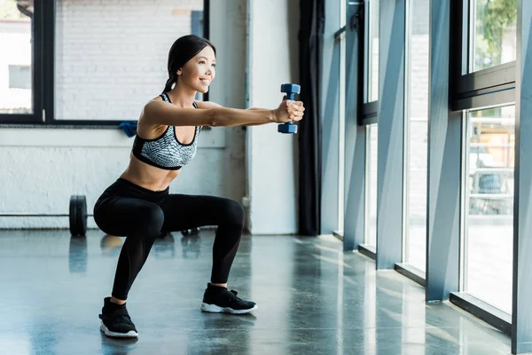 Happy young sportswoman holding dumbbells and doing squat exercise — Stock Photo