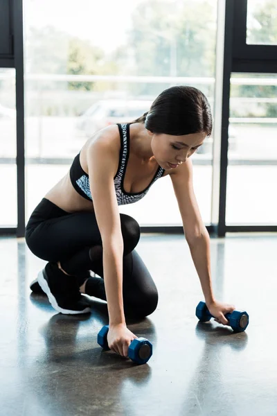 Young sporty woman taking dumbbells in gym near windows — Stock Photo