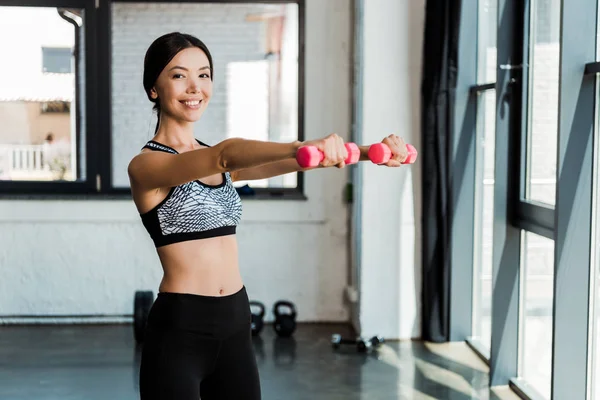 Chica feliz sosteniendo pesas rosadas mientras hace ejercicio en el gimnasio - foto de stock