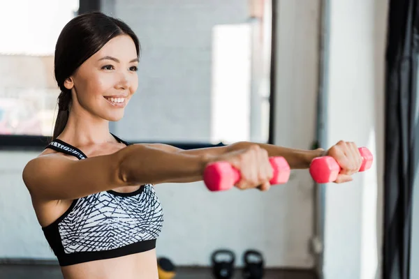 Enfoque selectivo de chica feliz sosteniendo pesas rosadas mientras hace ejercicio en el gimnasio - foto de stock