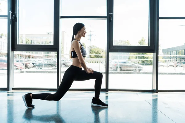 Vue latérale de la femme heureuse en vêtements de sport faire de l'exercice squat dans la salle de gym près des fenêtres — Photo de stock