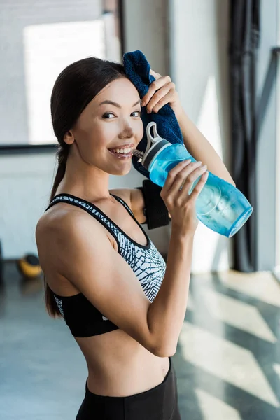 Веселая молодая женщина держит в руках спортивную бутылку и вытирает пот в спорткомплексе — стоковое фото