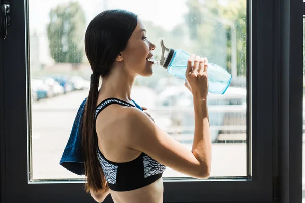 Vue latérale de fille heureuse eau potable tout en tenant la bouteille de sport dans la salle de gym — Photo de stock