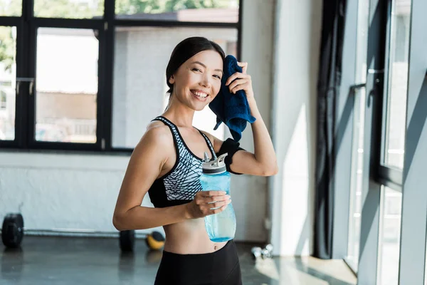 Chica feliz sosteniendo la botella de deporte mientras se limpia el sudor con toalla azul - foto de stock