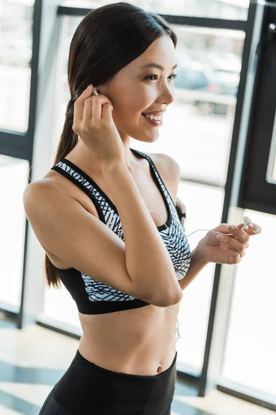 Mujer joven y deportiva feliz sosteniendo auriculares en el gimnasio - foto de stock