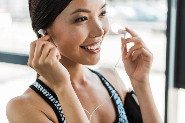 Primer plano de la mujer joven y deportiva feliz sosteniendo auriculares en el gimnasio - foto de stock