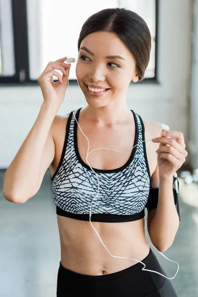 Mujer alegre y deportiva sosteniendo auriculares en el gimnasio - foto de stock