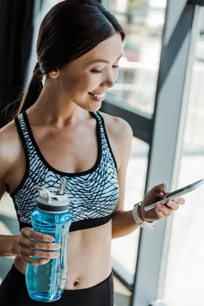 Jeune femme heureuse utilisant smartphone et tenant bouteille de sport — Photo de stock