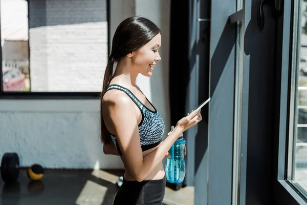 Vista lateral de la joven feliz usando el teléfono inteligente y la celebración de la botella deportiva - foto de stock