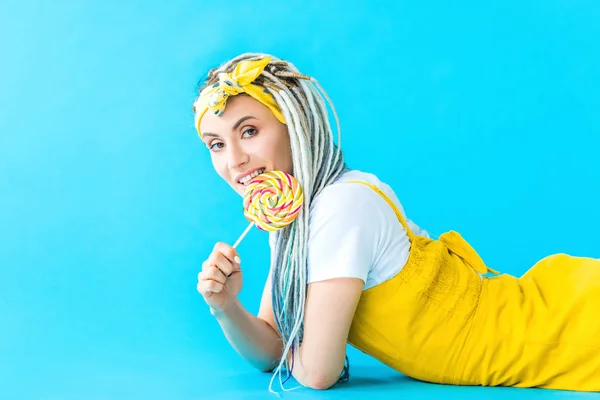 Smiling girl with dreadlocks lying and licking lollipop on turquoise — Stock Photo