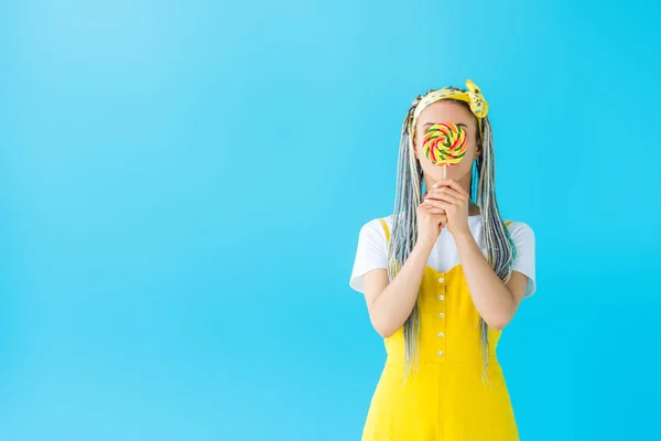 Girl with dreadlocks covering face with lollipop isolated on turquoise with copy space — Stock Photo
