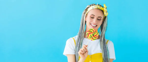 Panoramic shot of happy girl with dreadlocks holding lollipop isolated on turquoise — Stock Photo