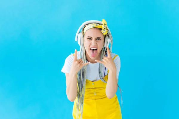Chica con rastas en los auriculares que sobresalen lengua y haciendo signos de roca aislados en turquesa - foto de stock