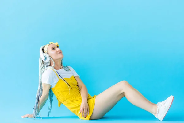 Chica con rastas en los auriculares sentado y mirando hacia arriba en turquesa - foto de stock