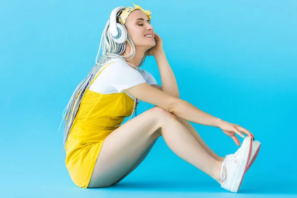 Fille souriante avec dreadlocks dans les écouteurs assis sur turquoise — Photo de stock