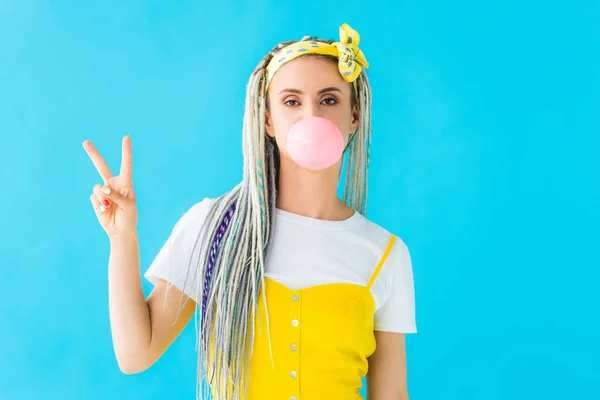 Chica con rastas soplando goma de mascar y mostrando signo de paz aislado en turquesa - foto de stock