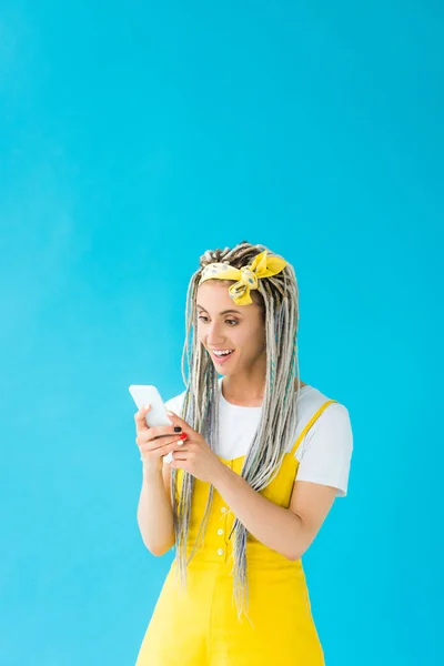 Chica feliz con rastas usando teléfono inteligente aislado en turquesa - foto de stock