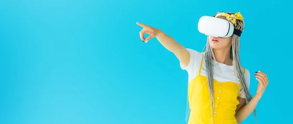 Plano panorámico de chica con rastas en auriculares de realidad virtual apuntando con el dedo aislado en turquesa - foto de stock