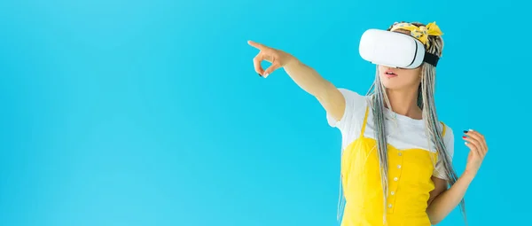 Panoramic shot of girl with dreadlocks in virtual reality headset pointing with finger isolated on turquoise — Stock Photo