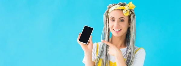 Панорамний знімок дівчини з дредлоками, що вказує пальцем на смартфон з порожнім екраном ізольовано на бірюзовому — стокове фото