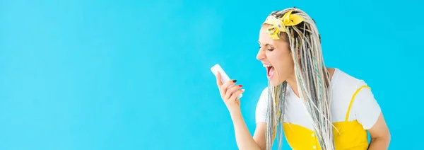 Panoramic shot of angry girl with dreadlocks yelling at smartphone isolated on turquoise — Stock Photo