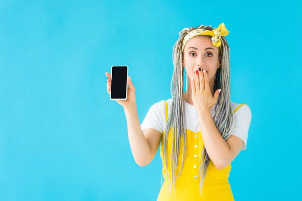 Дівчина з дредлоками, що закривають рот і показують смартфон з порожнім екраном ізольовано на бірюзовому — стокове фото