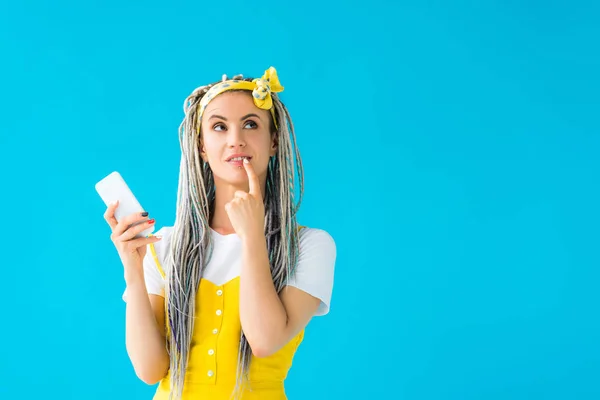 Chica pensativa con rastas y dedo en la boca sosteniendo teléfono inteligente aislado en turquesa - foto de stock