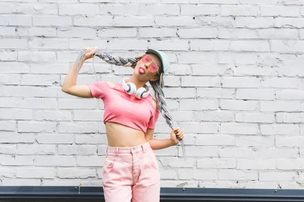 Hermosa chica en sombrero con auriculares que sobresalen lengua cerca de la pared de ladrillo - foto de stock